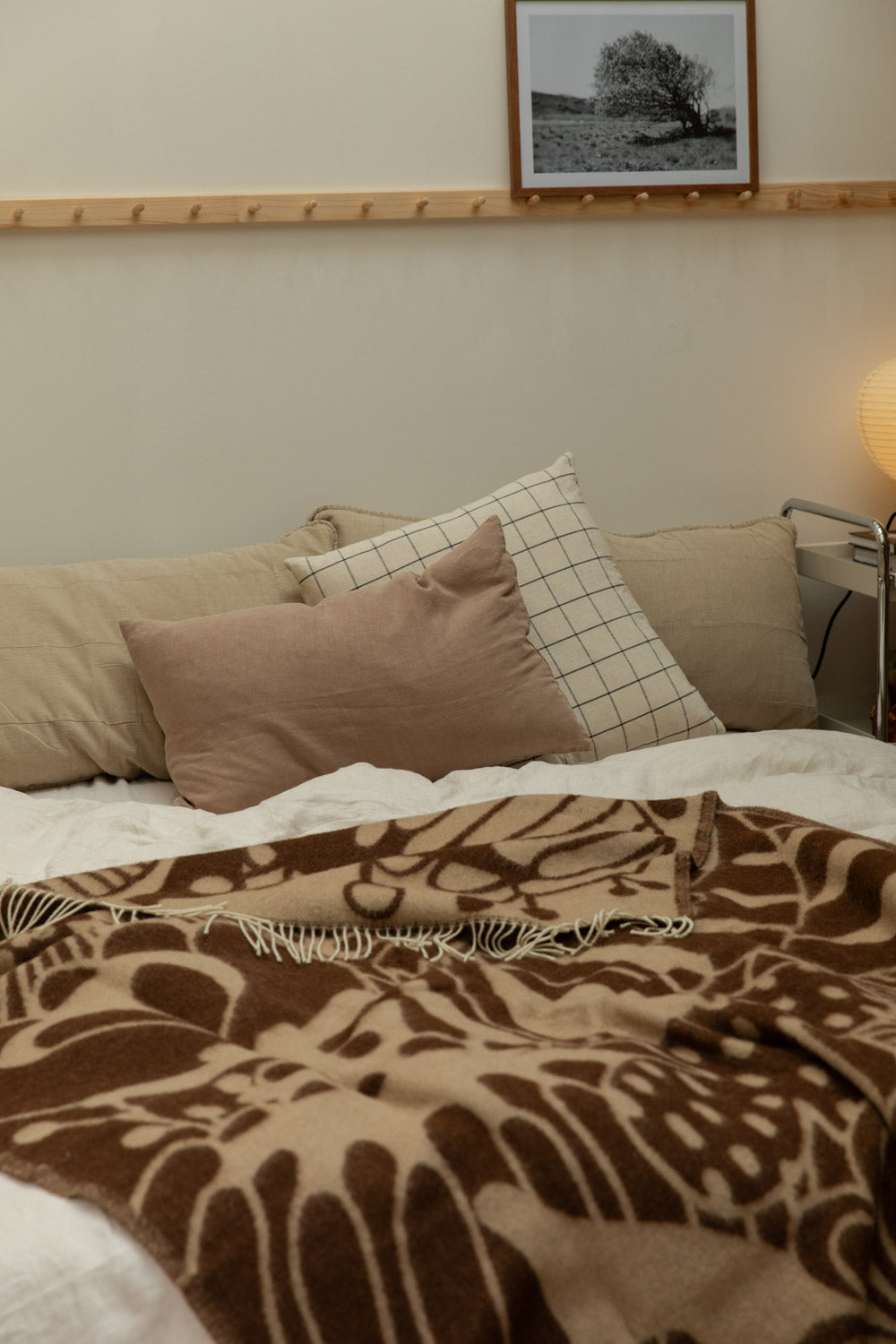 Klippan Blomranka lambswool blanket in chocolate brown and sand draped over a bed with pine peg rail, black and white tree art print and glowing paper lantern table lamp.