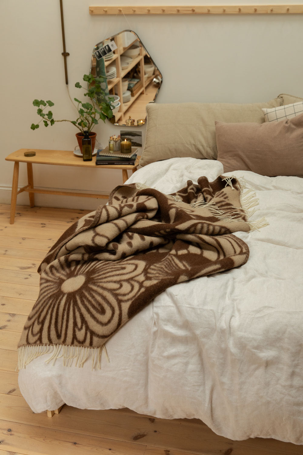 Klippan Blomranka lambswool blanket in chocolate brown and sand casually draped over end of bed with organic mirror, trailing plant, lit candle and pine bench bedside table.