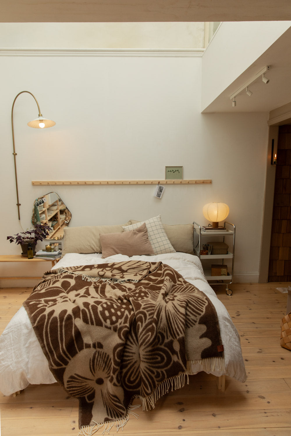 Full bedroom view with Klippan Blomranka lambswool blanket in chocolate brown and sand draped over a bed with arc brass floor lamp, organic mirror and pine floorboards.