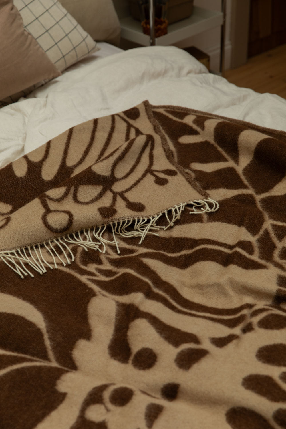Close-up of Klippan Blomranka lambswool blanket in chocolate brown and sand showing bold floral print and cream fringe edge detail draped over a bed.