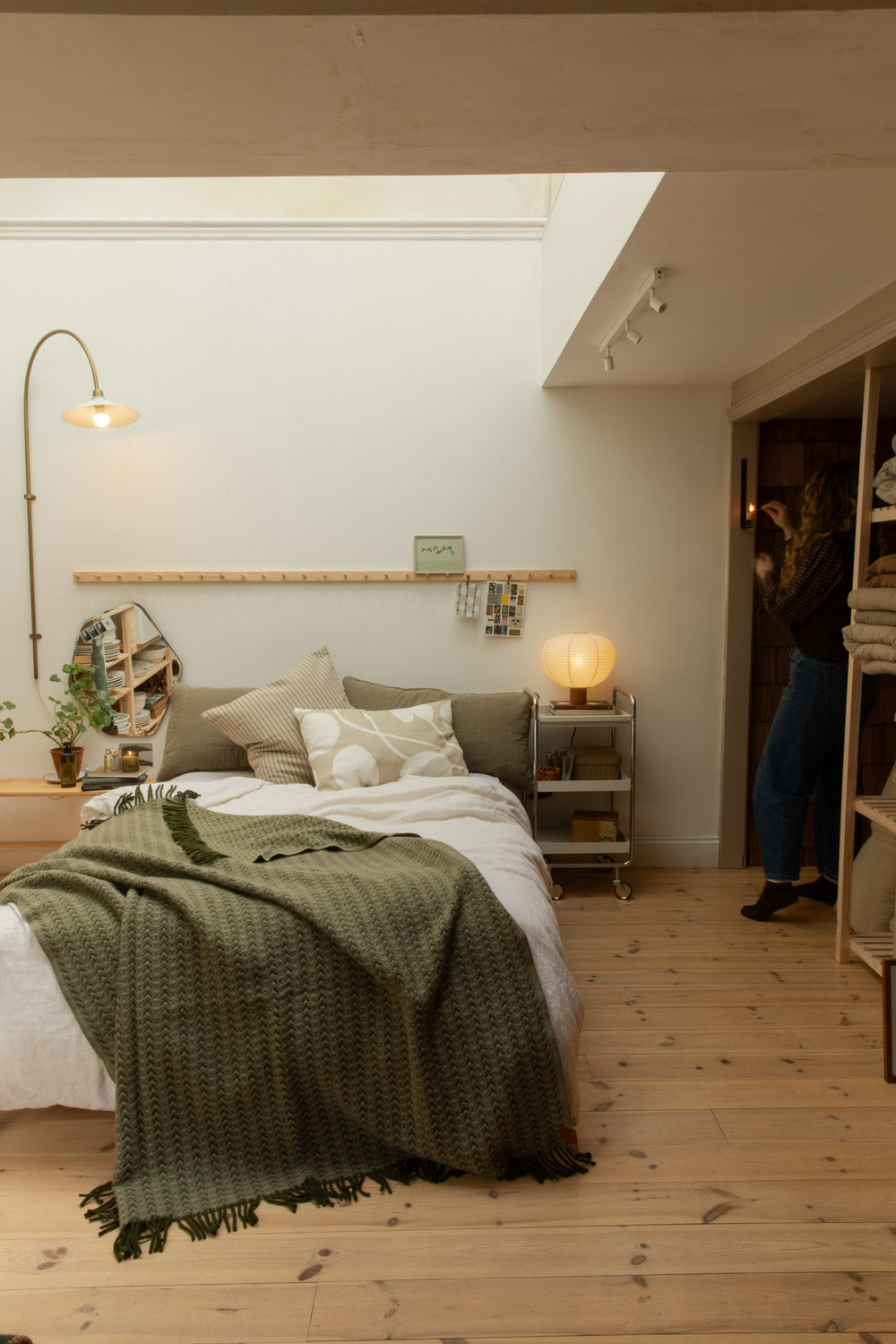 Full bedroom lifestyle shot with Ilse Wood olive green wool blanket draped on white linen bed, Blomus Kami lamp, brass floor lamp and pine peg rail
