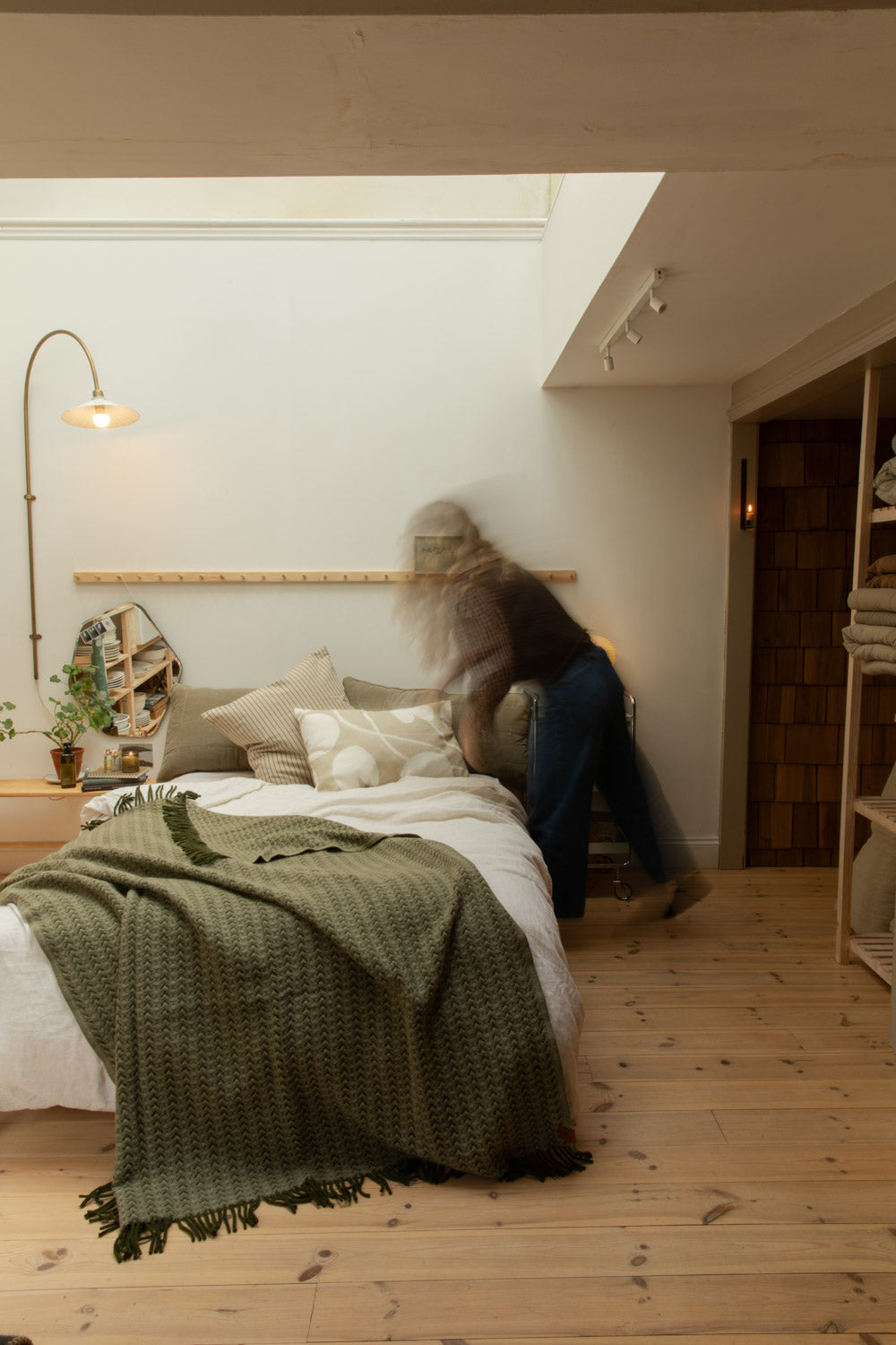 Motion blur lifestyle shot of bedroom with Ilse Wood olive green wool blanket on white linen bed, Blomus Kami lamp and brass floor lamp.