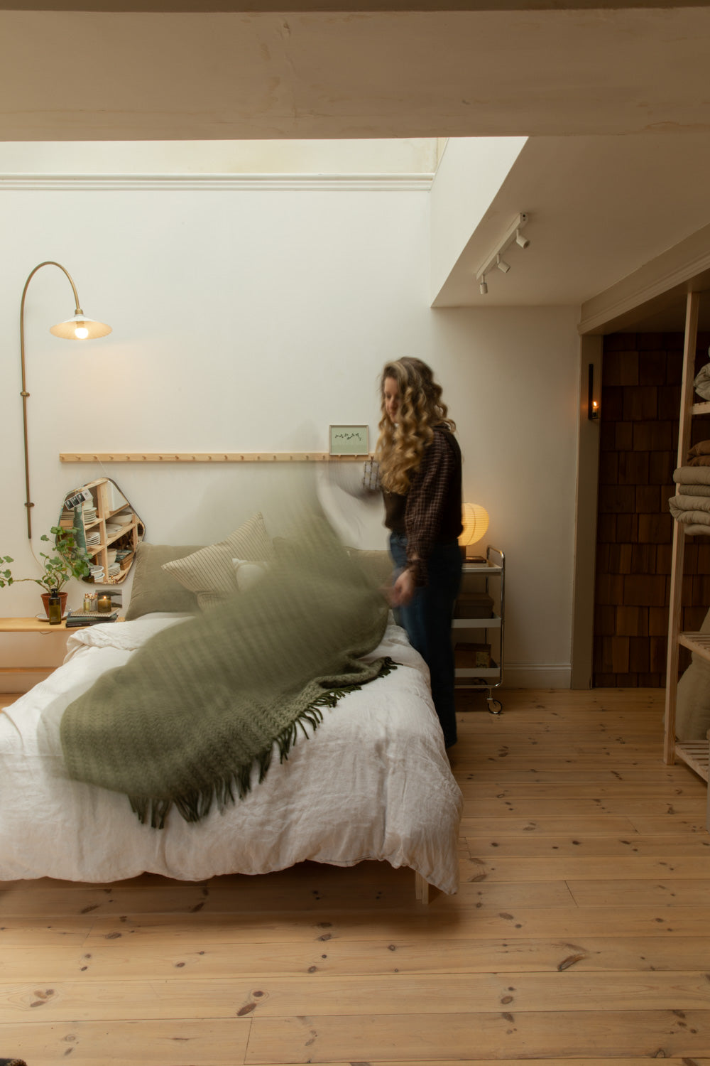 Woman draping Ilse Wood olive green wool blanket across a white linen bed, Blomus Kami table lamp and brass floor lamp in warm bedroom setting.