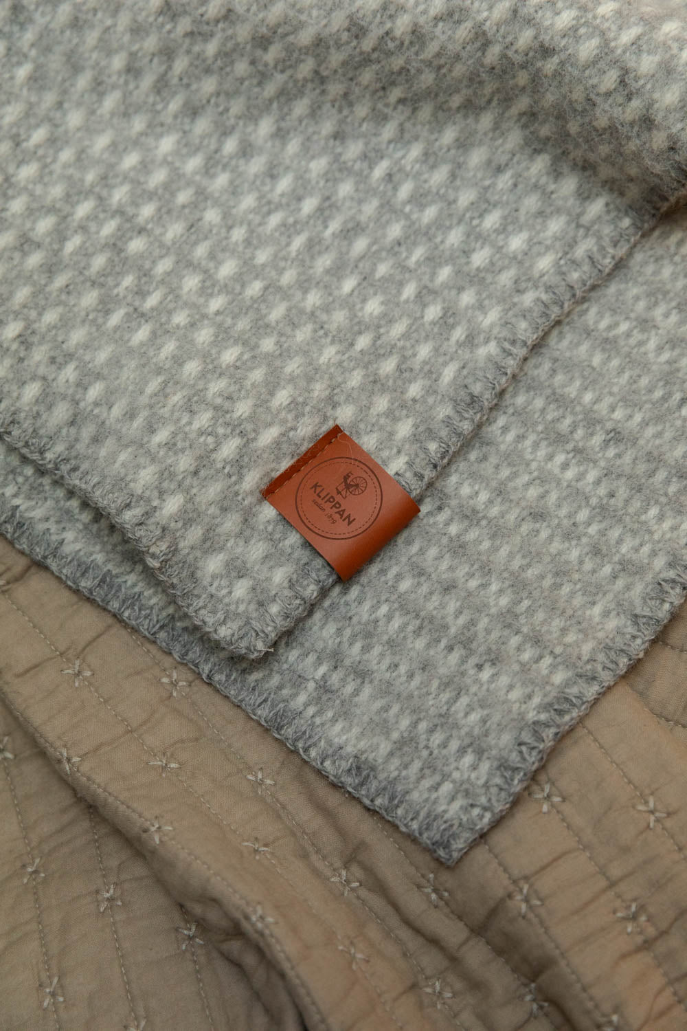 Close detail of Klippan leather label on grey and white waffle weave lambswool baby blanket, layered over a vintage linen cross stitch quilt.