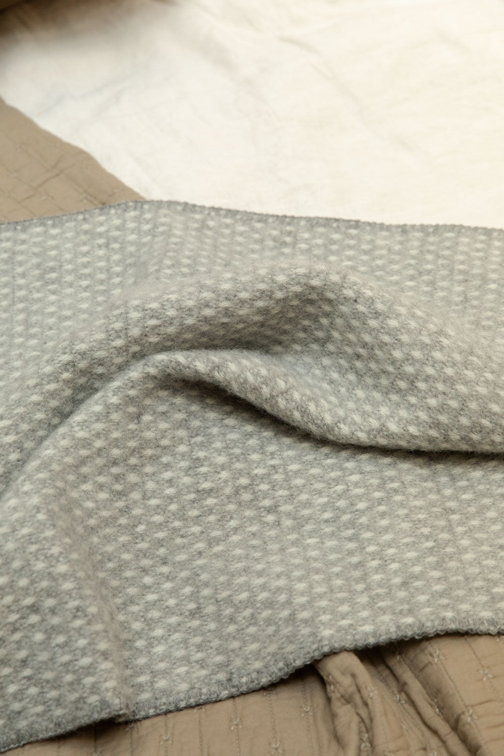 Close detail of Klippan Knut lambswool baby blanket in grey and white waffle weave, draped over a cotton cross stitch quilt on a white linen bed.