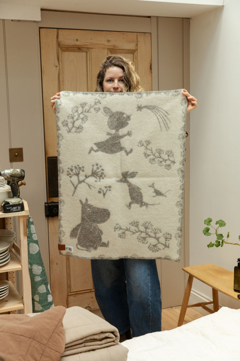 Klippan Moomin recycled lambswool baby blanket in cream held up in shop interior showing full reversible design with Moomintroll, Little My and Sniff characters.