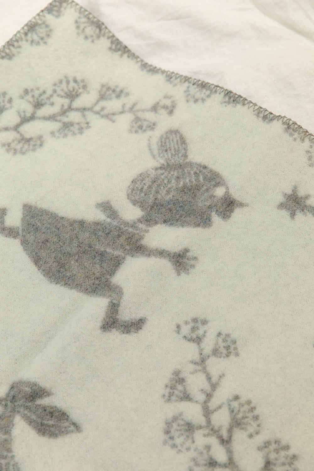 Close-up of Klippan Moomin recycled lambswool baby blanket in grey and cream showing Little My character and botanical print detail on cream side.