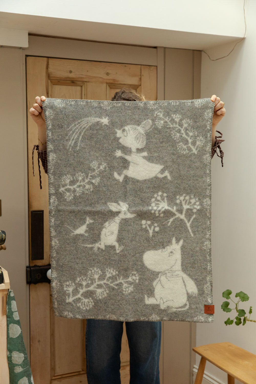 Klippan Moomin recycled lambswool baby blanket in grey held up showing full design with Moomintroll, Little My and Sniff characters among botanical branches.