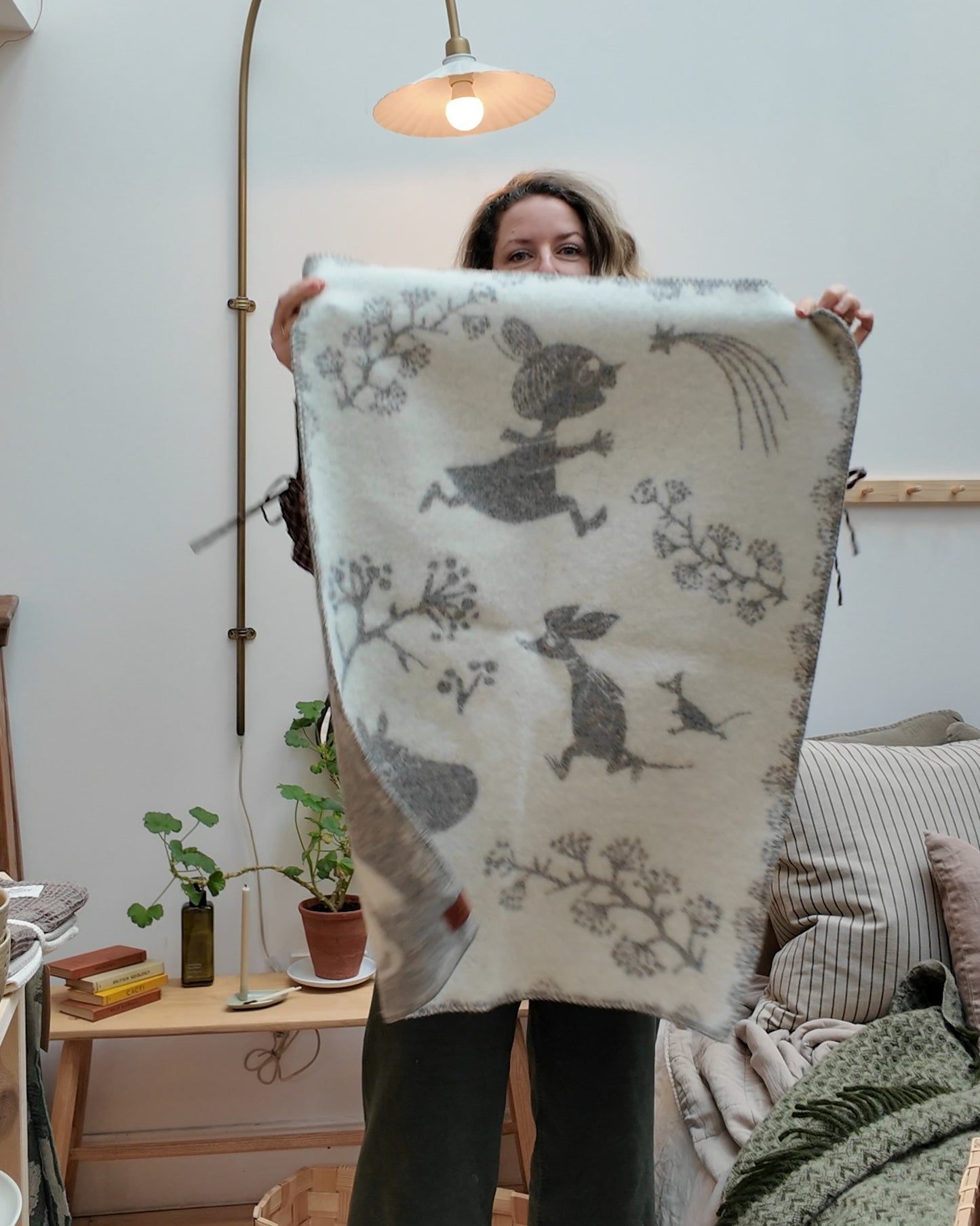 Scandinavian styled room with Alice holding up a white and grey moomin Children’s wool blanket.