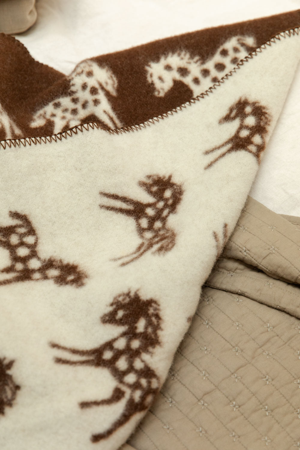 Klippan Pony lambswool mini blanket in cream with brown horse print, draped over a bed with a vintage linen cotton quilt.