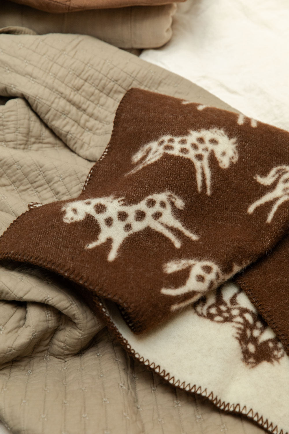 Klippan Pony lambswool mini blanket showing chocolate brown reverse with cream horse print, folded on a bed with vintage linen cotton quilt.