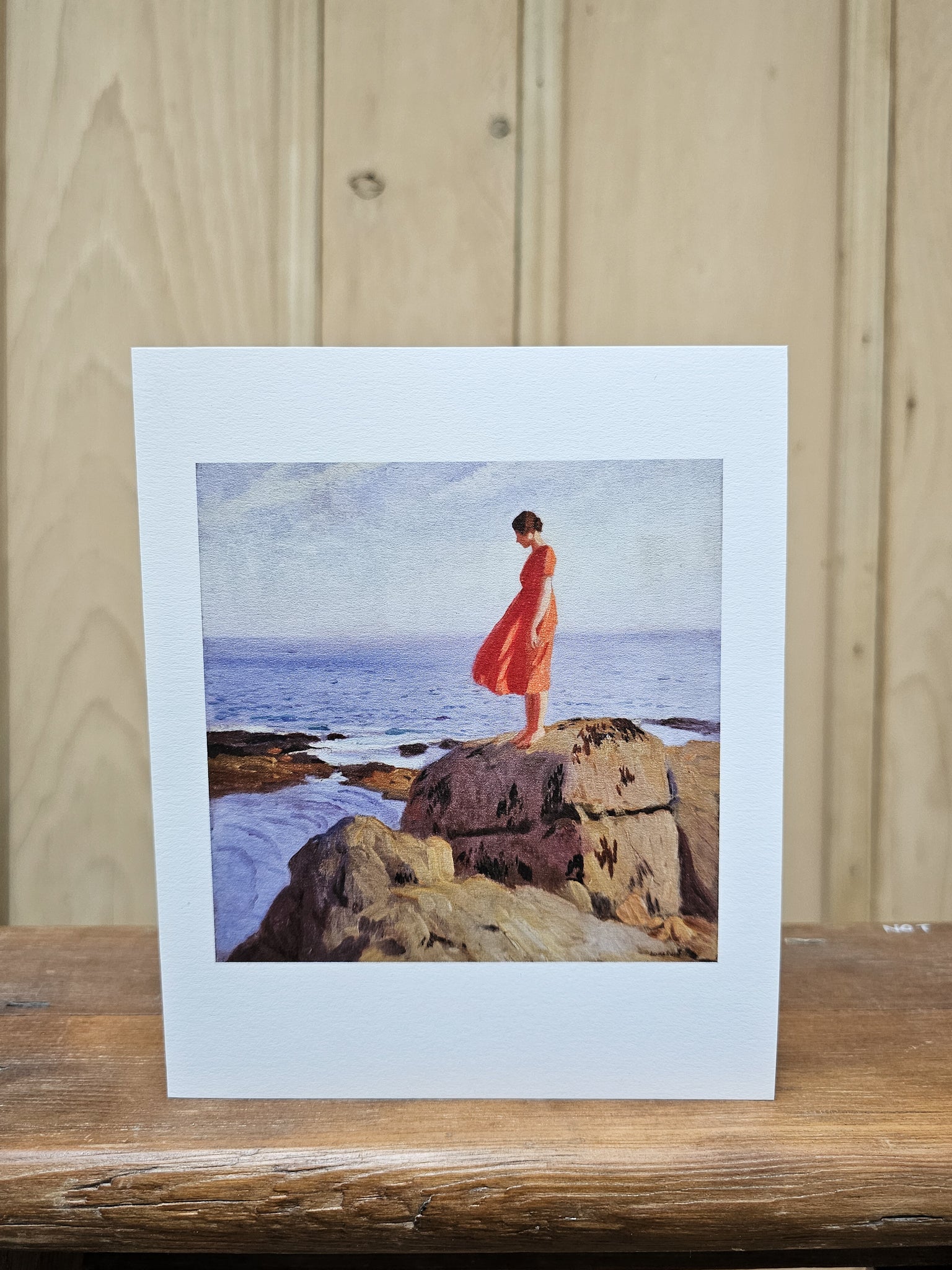 Greeting card reproducing Laura Knight's oil painting "A Dark Pool" with a woman in a flowing red dress standing barefoot on coastal rocks, gazing down towards a calm blue sea