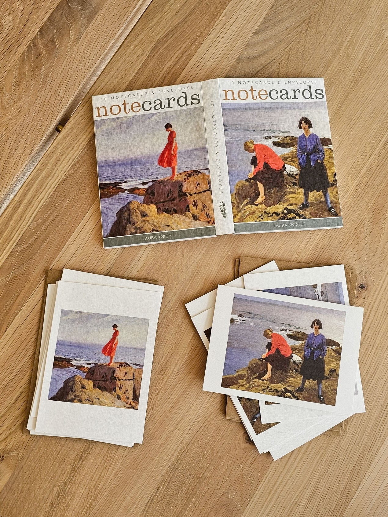 A set of illustrated Laura Knight art notecards arranged on a light wood tabletop. The open pack at the top shows the title 'notecards' and a coastal scene with people standing on rocks; several cards are stacked in front, showing the same print design.