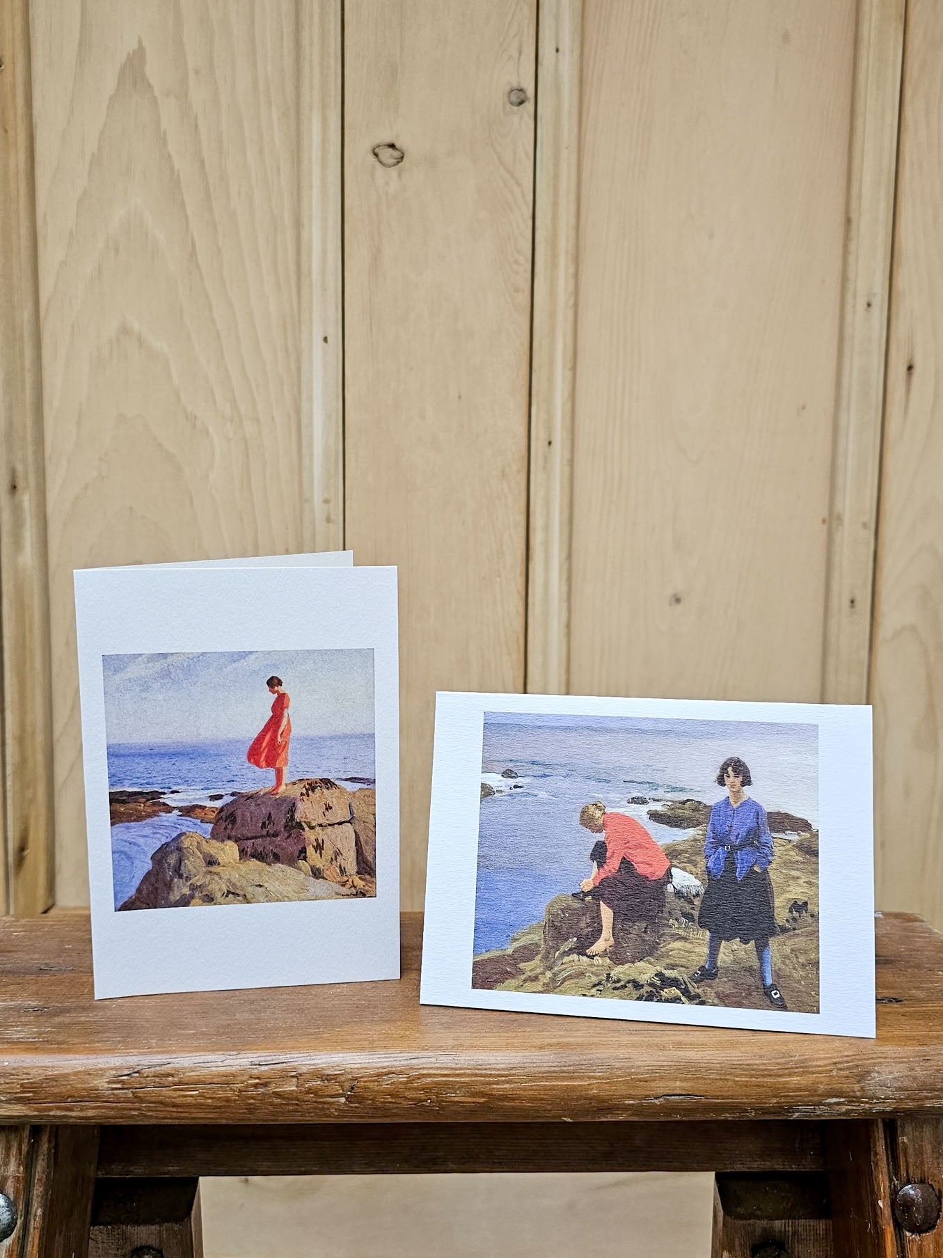 Two art prints featuring a coastal scene with figures on rocks, displayed on a rustic wooden table against a light wood paneled wall.