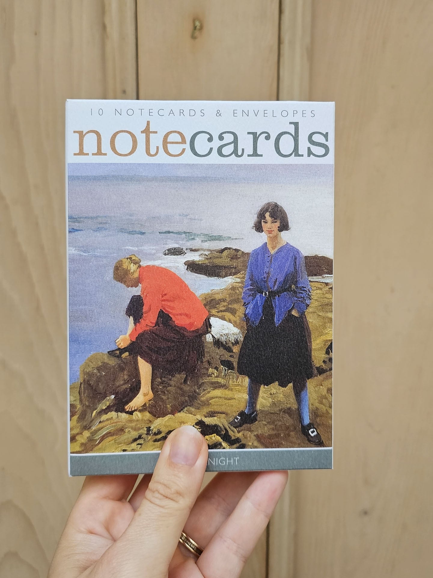A hand holds a boxed set of 'notecards' against a light wood background. The box features an illustration of two women on a rocky coastline, with the title 'notecards' printed at the top.