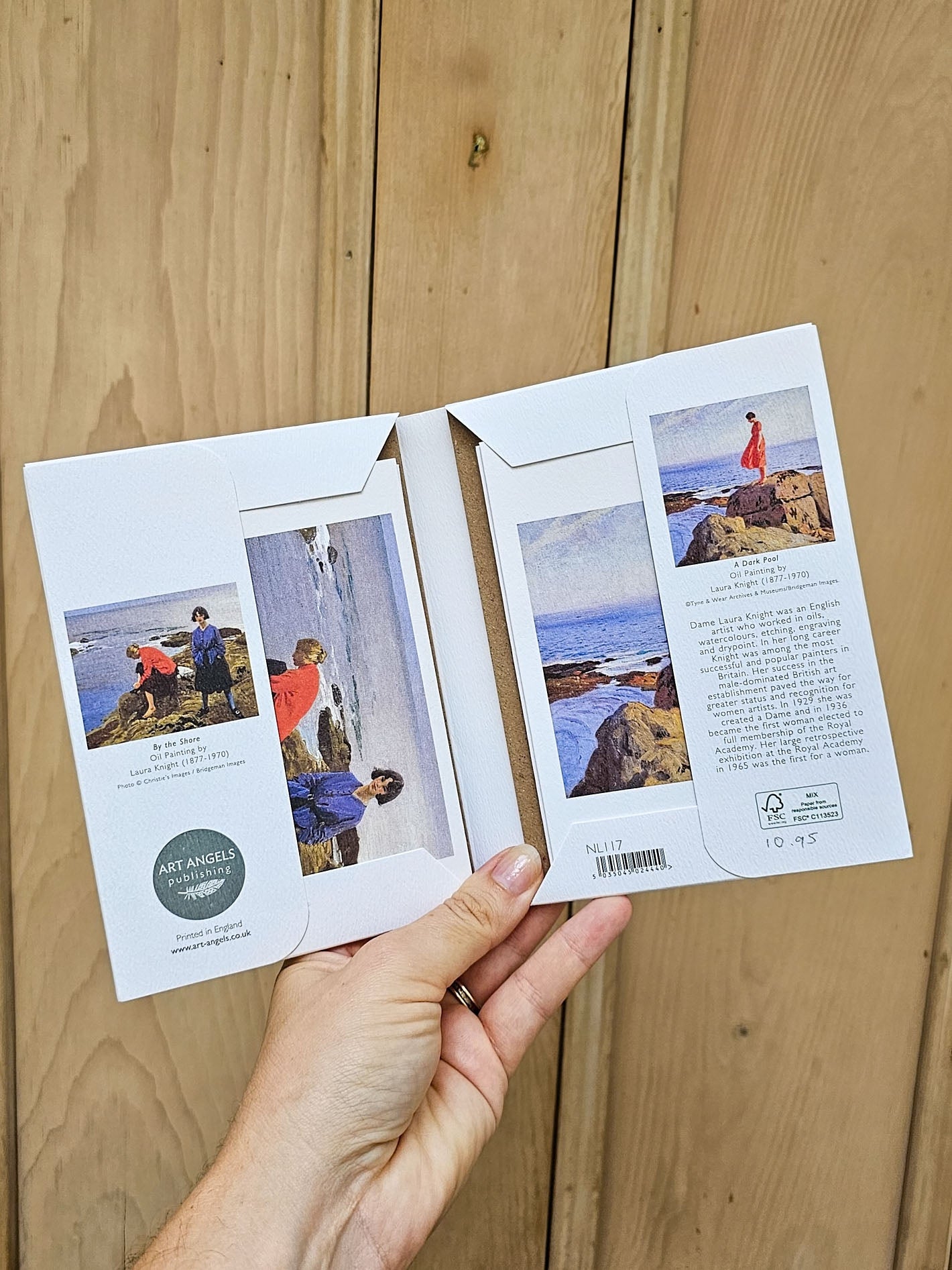 A hand holds an open pack of notecards light wood plank background. Inside are printed cards with artwork of coastal scenes with people and descriptive text, plus a circular green seal reading Art Angels London.