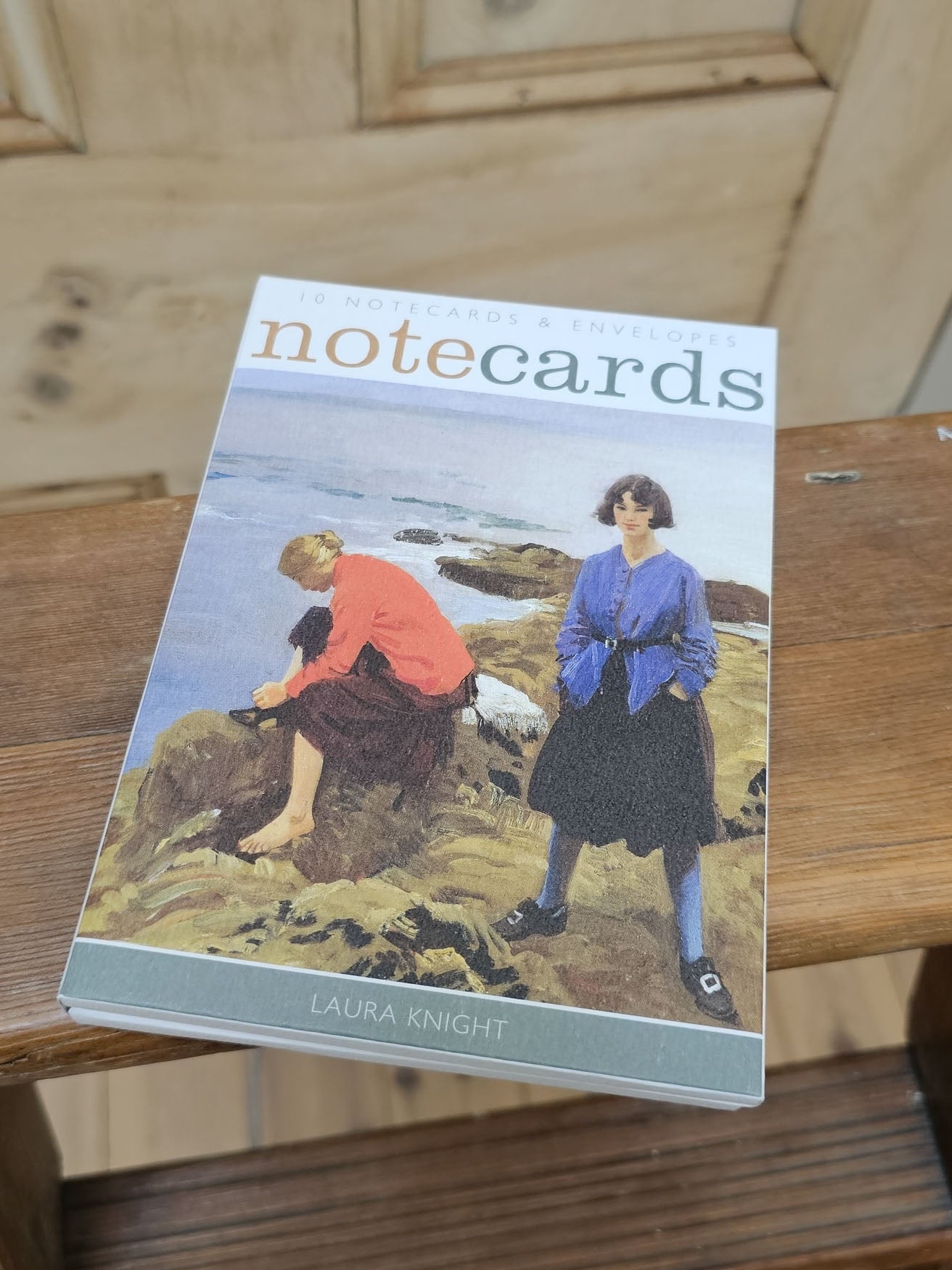 Notecards by Laura Knight resting on a wooden surface. The cover features an illustration of two women on a rocky beach, with the title 'notecards' in a muted orange-green font.