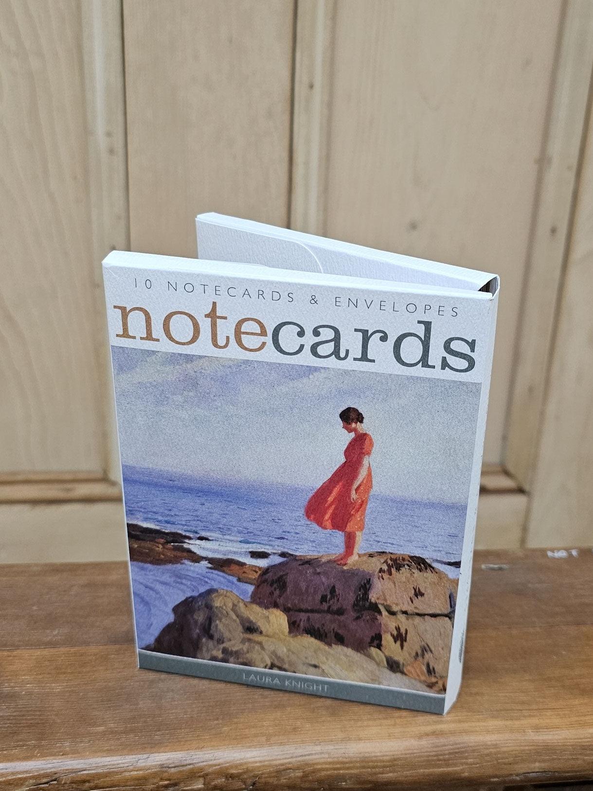 A boxed set of notecards and envelopes standing upright on a wooden surface against a light wood paneled background. The front cover shows the title 'notecards' and an illustration of a person in a red dress standing on rocks by the sea.