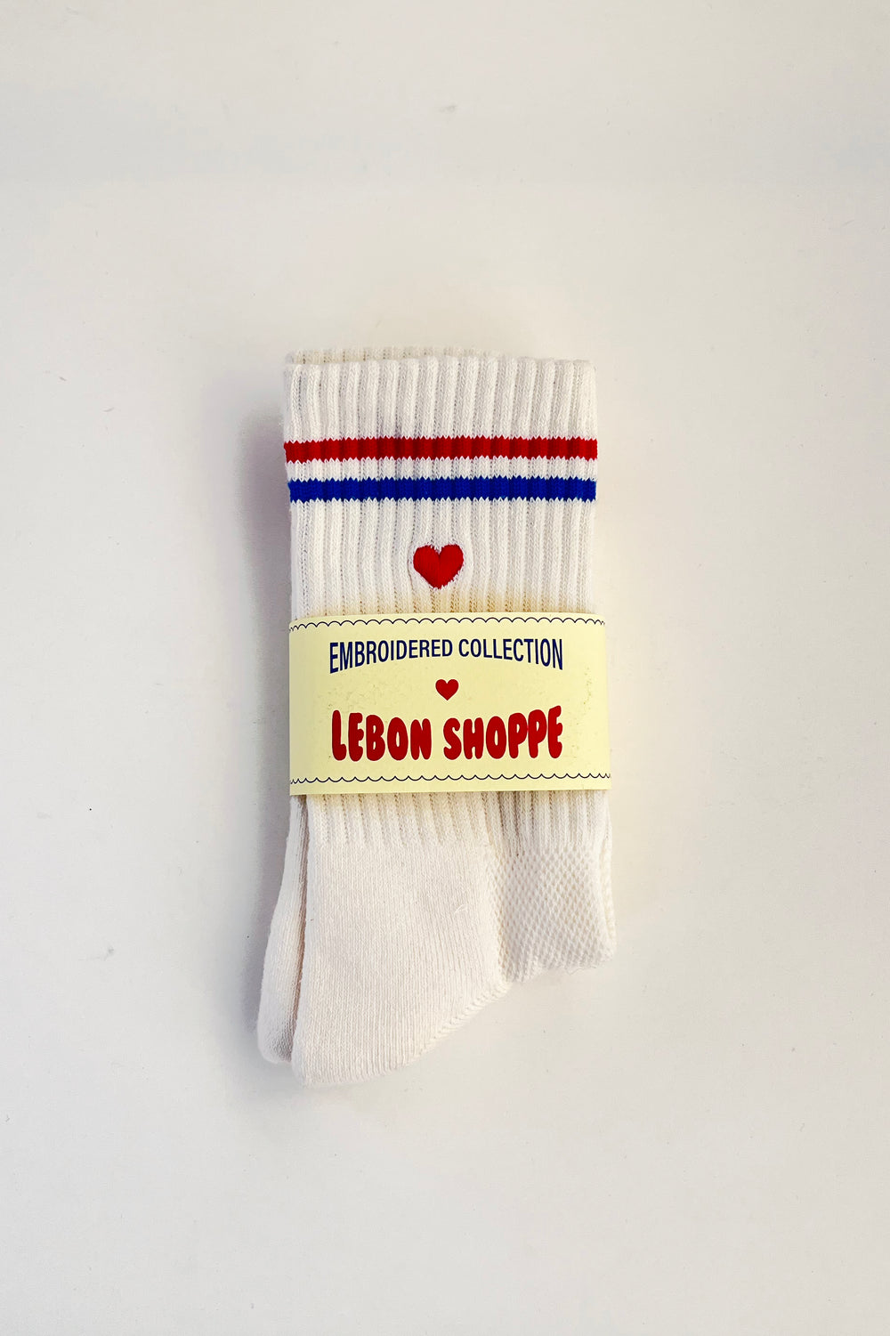 White sock with redand blue stripes and an embroidered red heart folded in its Le Bon Shoppe packaging against a white background.