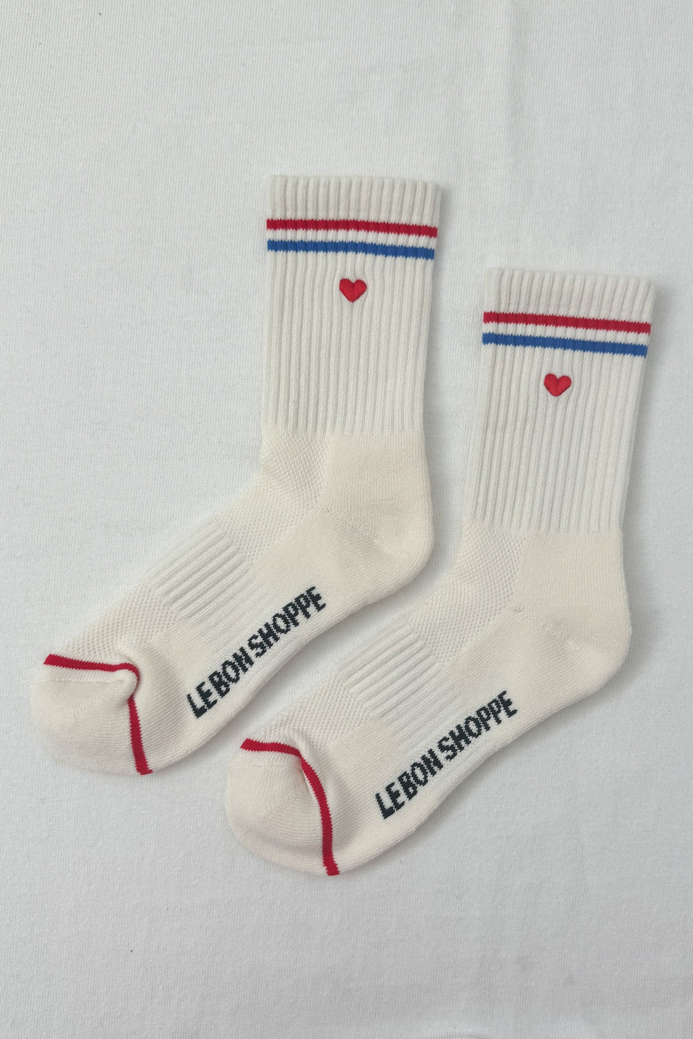 Pair of Le Bon Shoppe Boyfriend Socks in white with red and blue stripe and embroidered heart detail, flat lay.