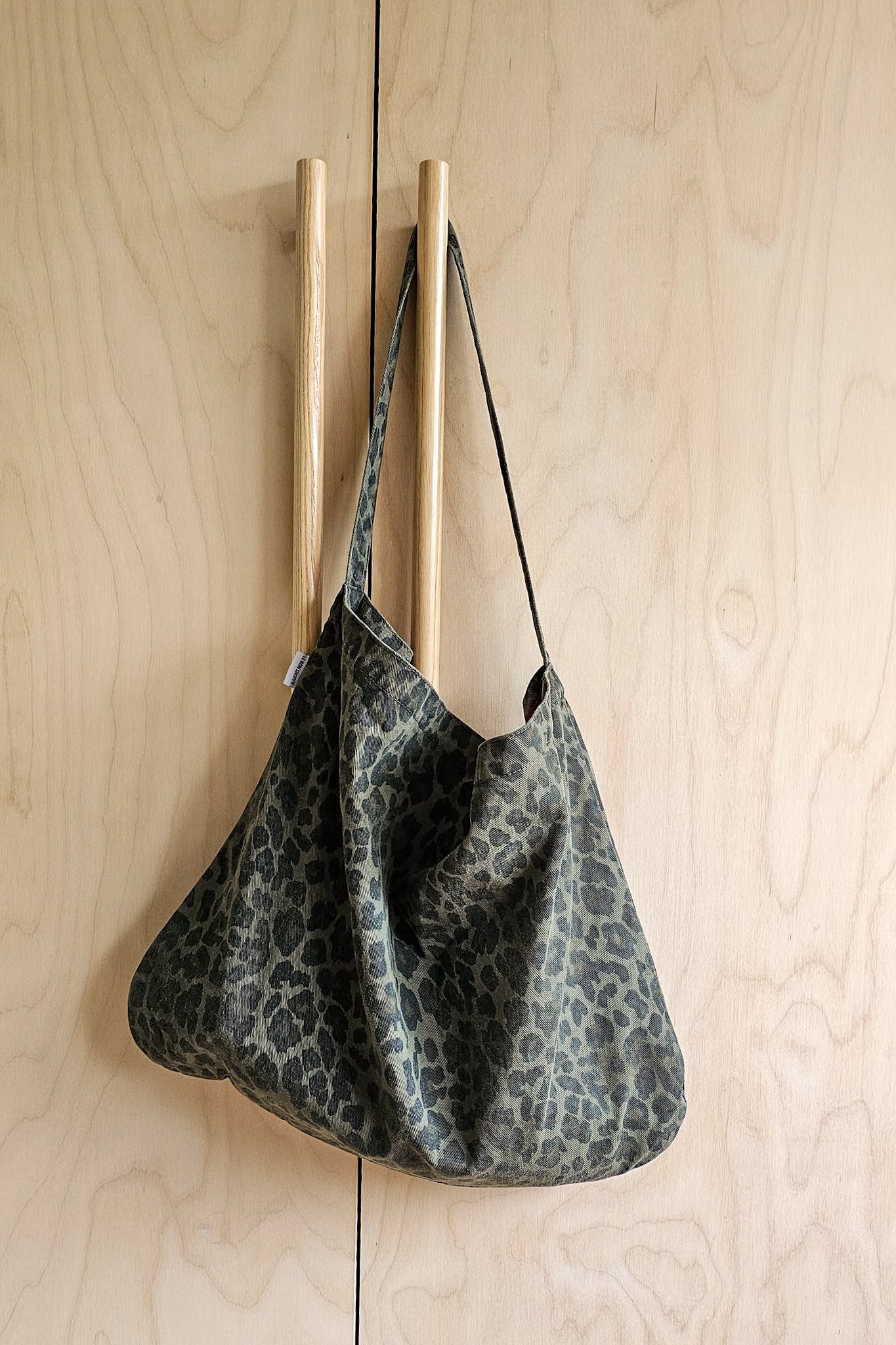 Large slouchy leopard print canvas tote bag in muted khaki green, hung on wooden peg rail against plywood wall.