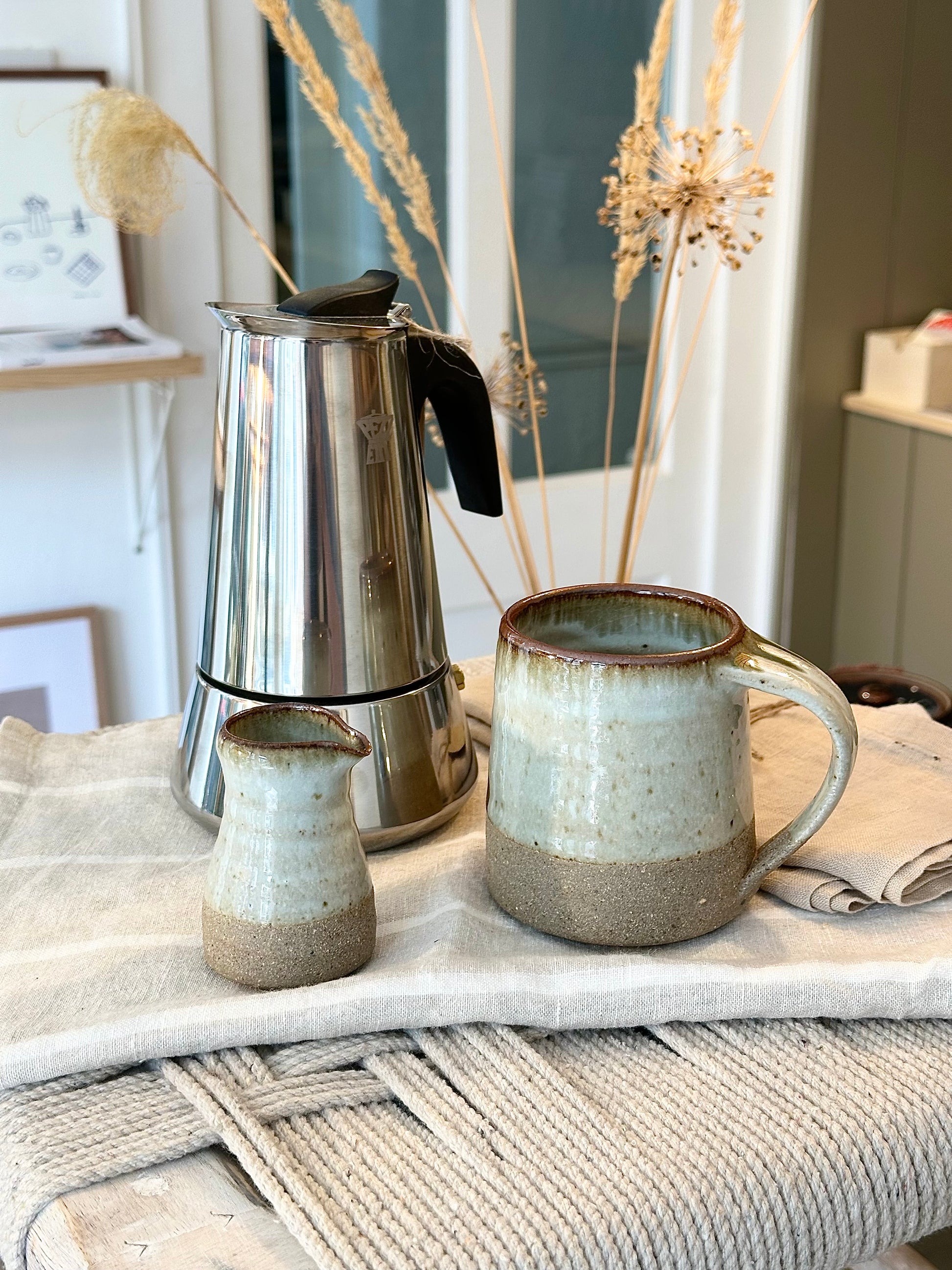 A Leach Pottery stoneware pourer with a 100ml capacity, displayed alongside a silver moka coffee maker and a Leach mug, on a white textured surface.