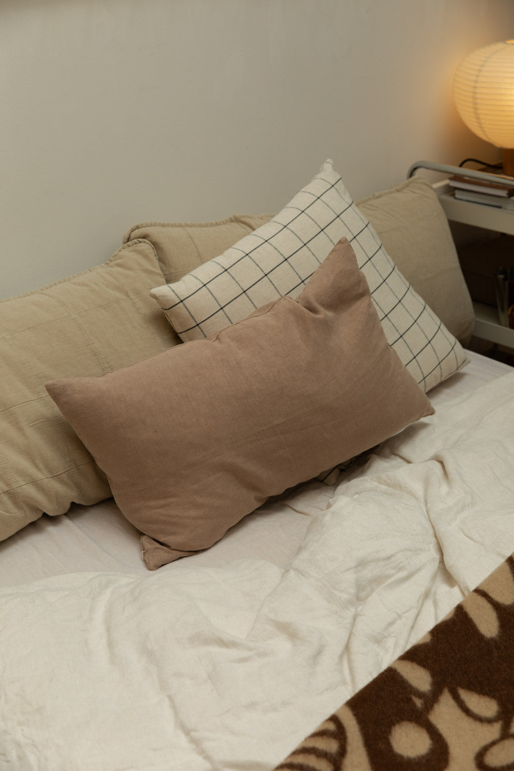 Cognac linen cushion cover layered with a grid print cushion on a white linen bed, Blomus Kami lamp glowing on chrome bedside trolley.