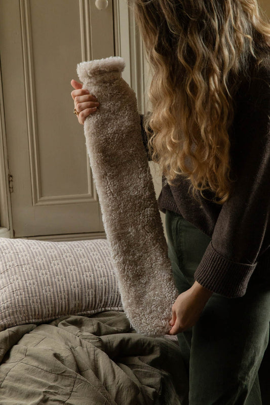 Woman holding an extra long sheepskin hot water bottle in natural oatmeal beside a quilted window seat
