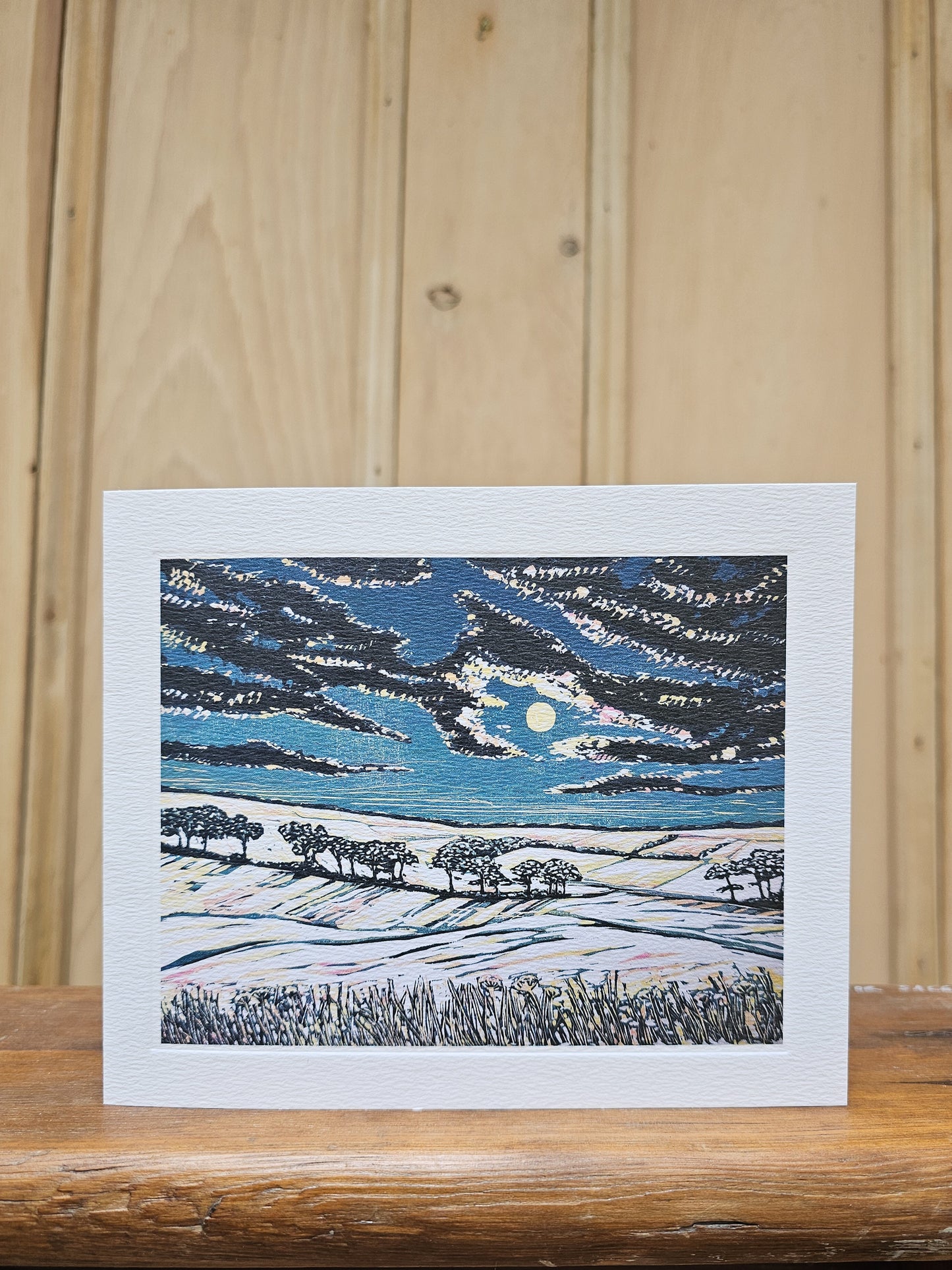 Greeting card reproducing Louise Stebbing's linocut print Full Moon, showing a pale moon breaking through dramatic dark clouds over a snowy hillside with silhouetted trees