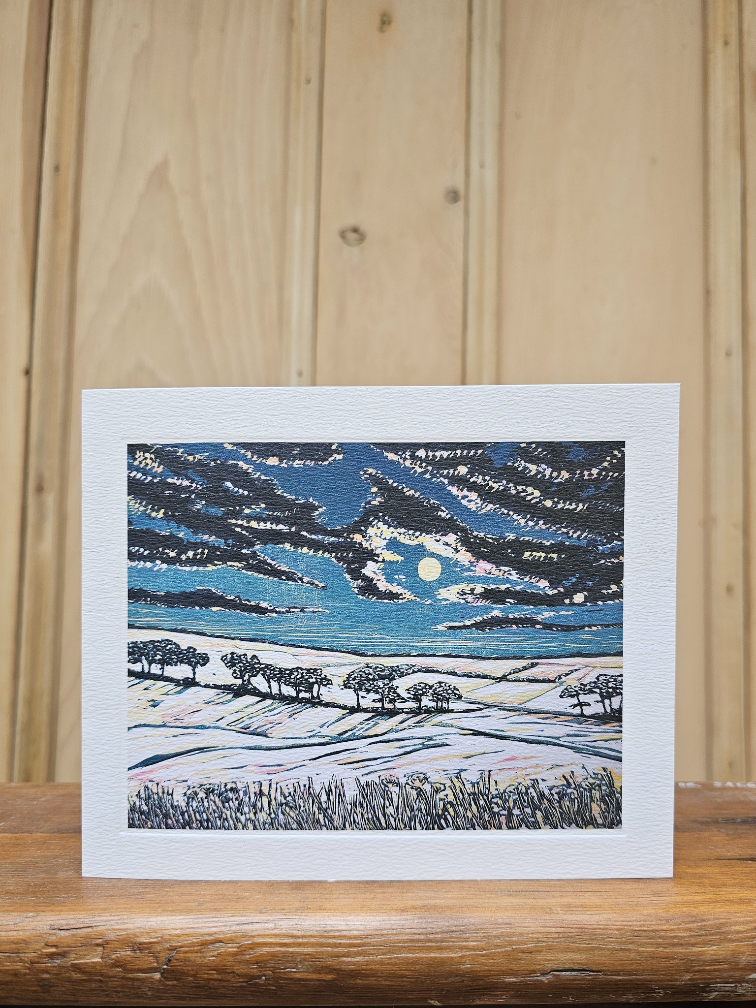 Greeting card reproducing Louise Stebbing's linocut print Full Moon, showing a pale moon breaking through dramatic dark clouds over a snowy hillside with silhouetted trees