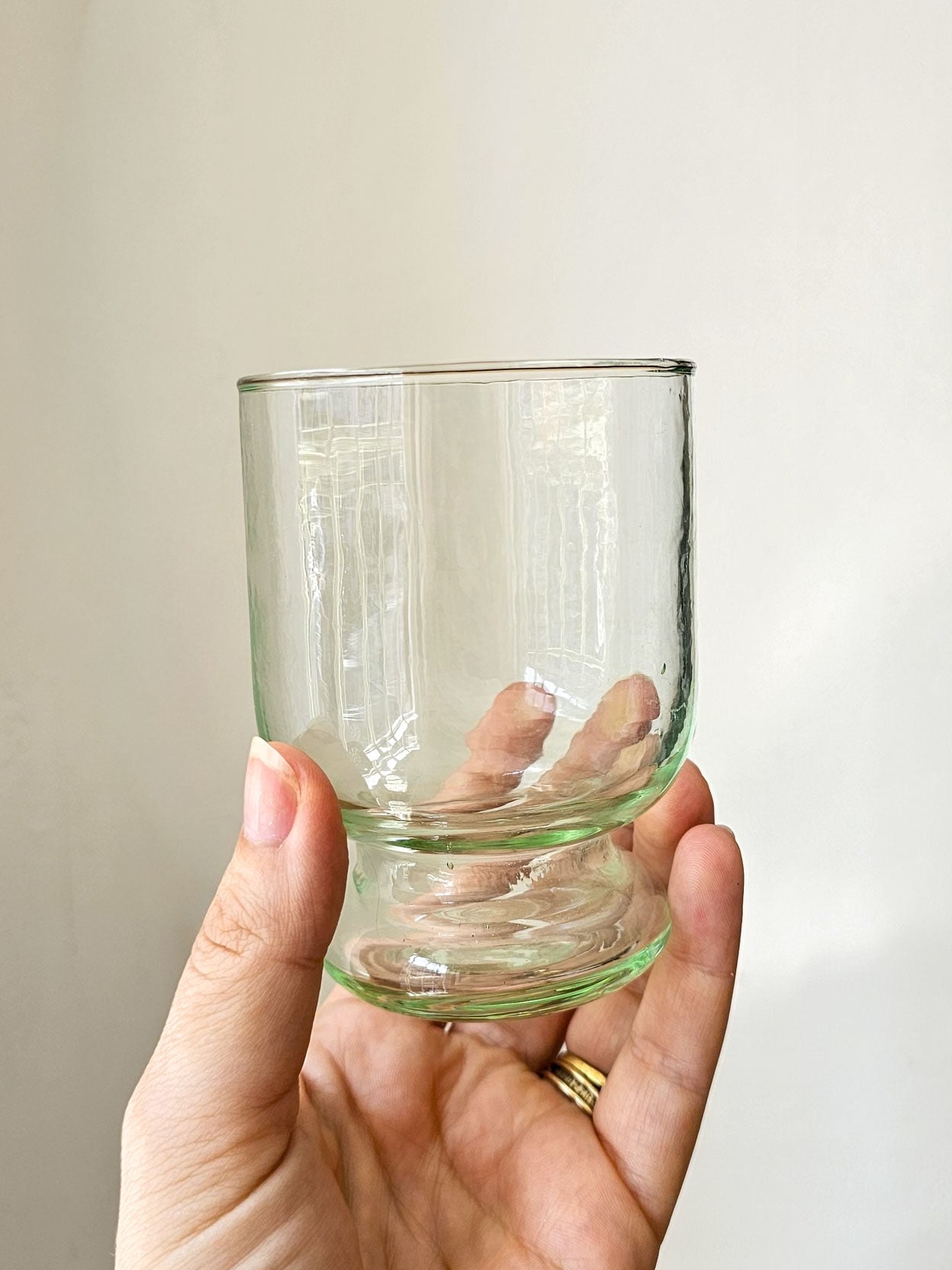 A hand holding a clear, light green-tinted glass tumbler against a plain, light beige background.