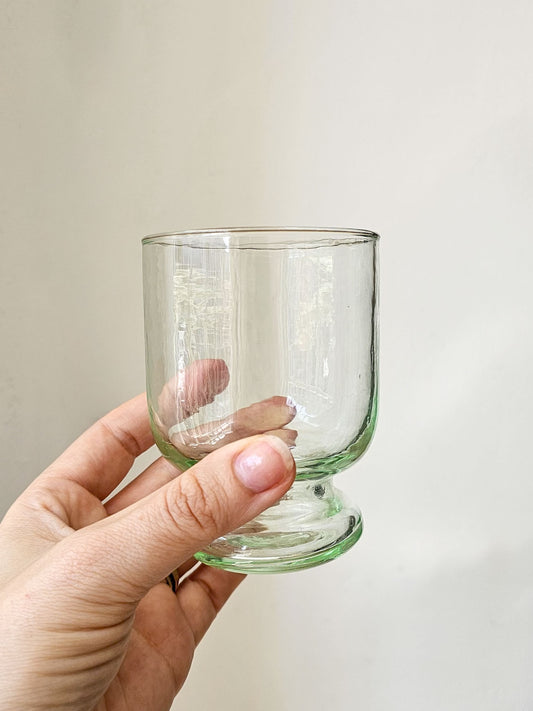 A hand holding a small clear glass tumbler with a light green tinted rim and base against a plain off-white background.