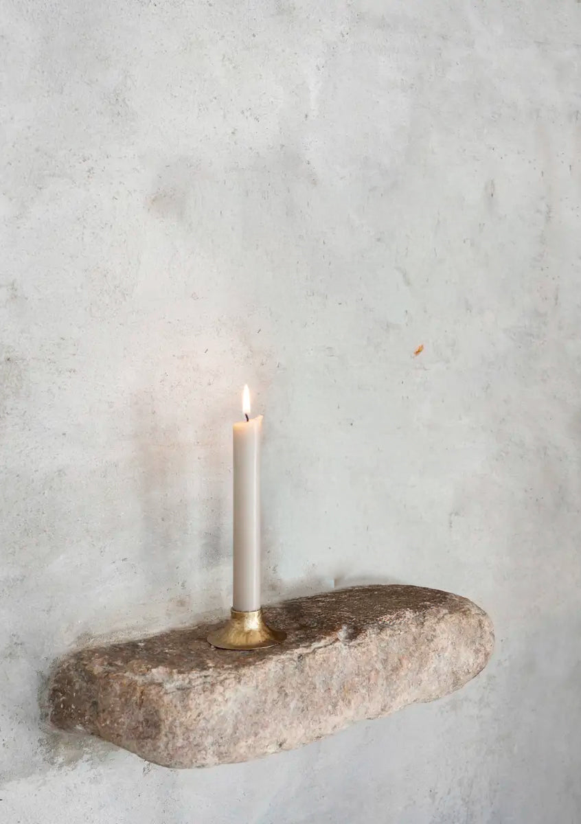 A lit white taper candle in a brass holder set into a rough stone slab mounted on a textured light gray plaster wall.
