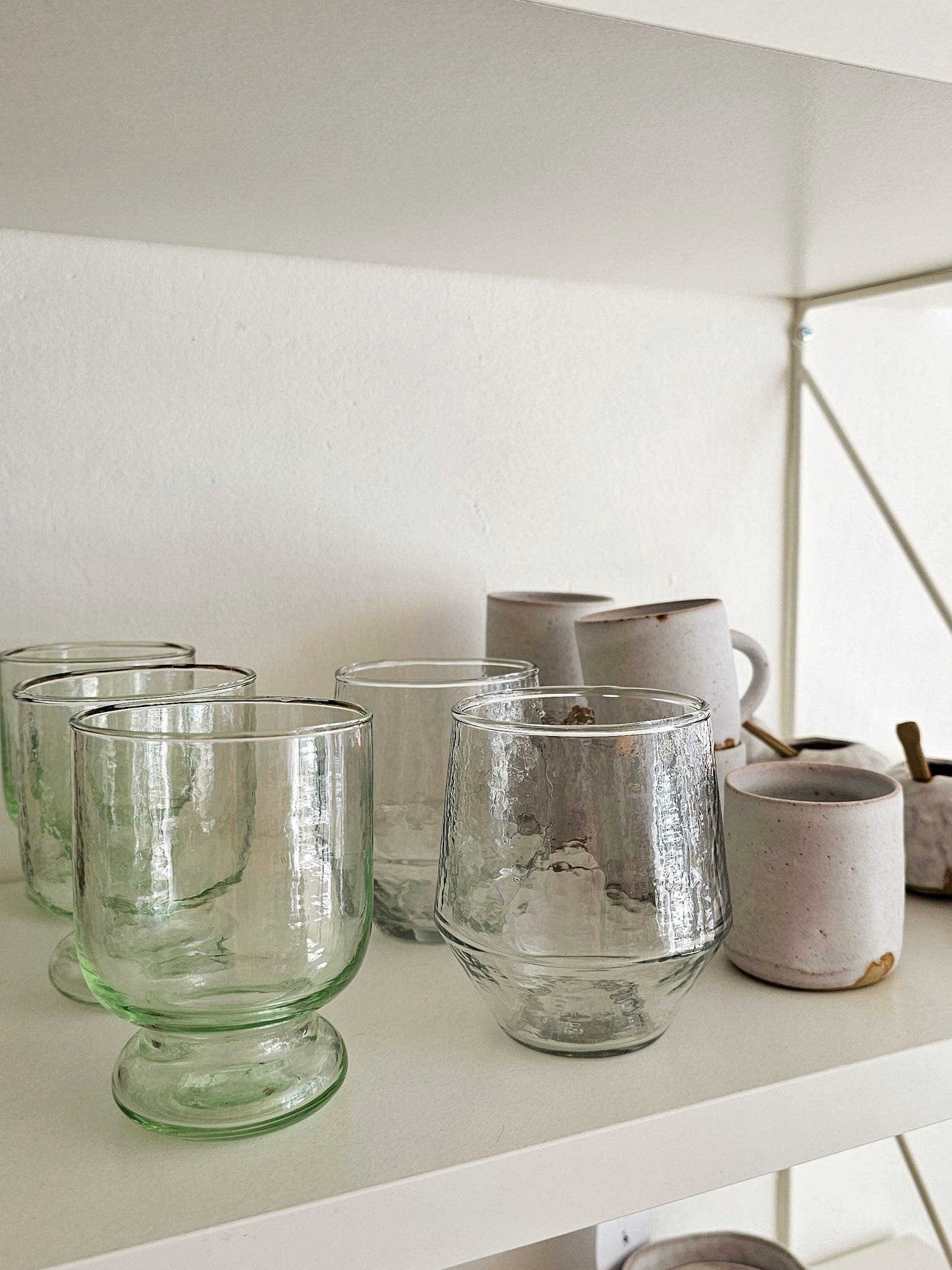 A set of clear glass drinking glasses and tumblers displayed on a white shelf alongside light gray ceramic mugs and small bowls.