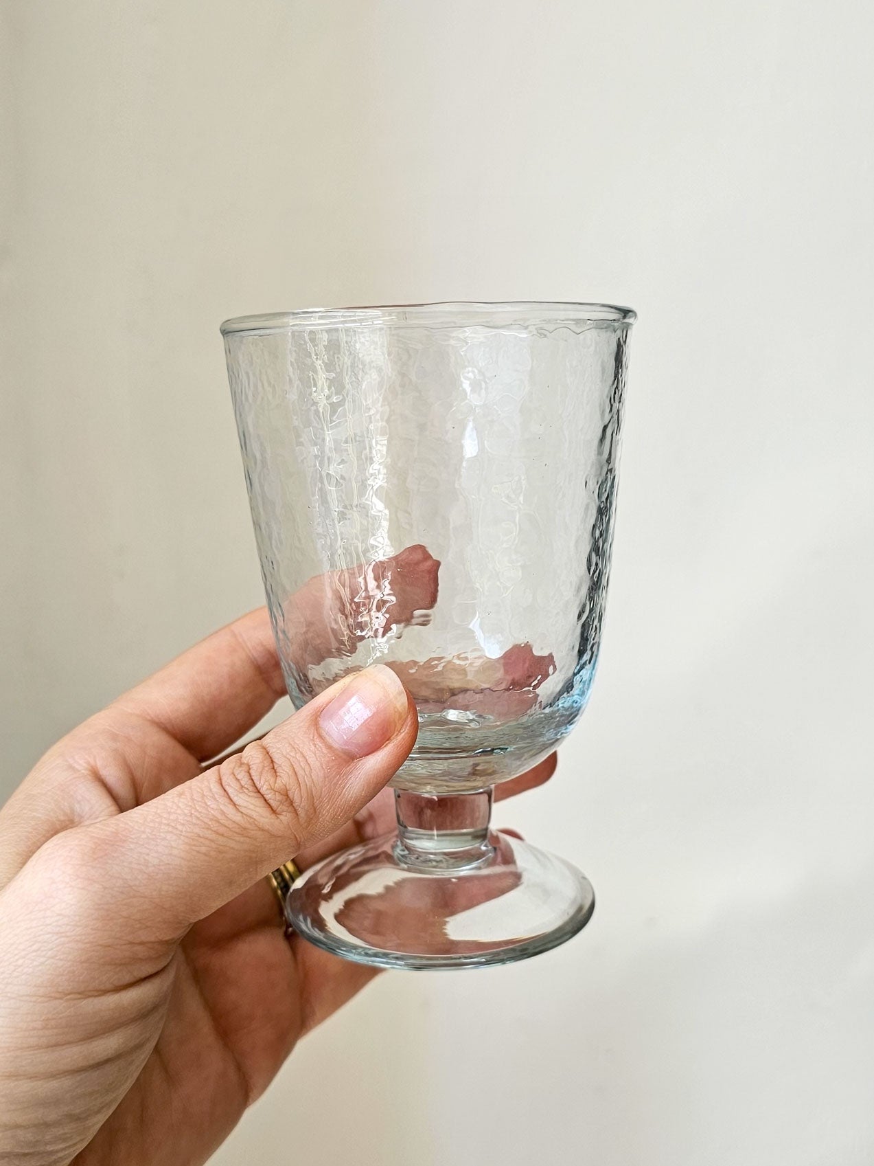 A hand holding a clear glass goblet with a hammered texture and a short stem against a plain light beige background.