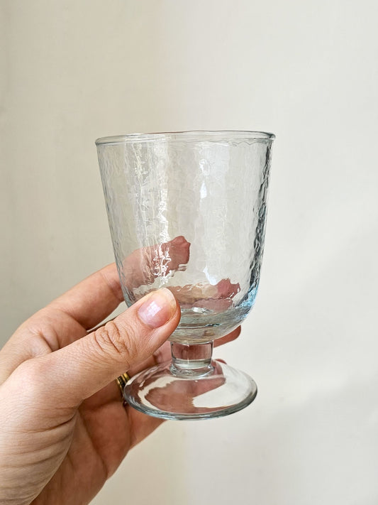 A hand holding a clear glass goblet with a hammered texture and a short stem against a plain light beige background.