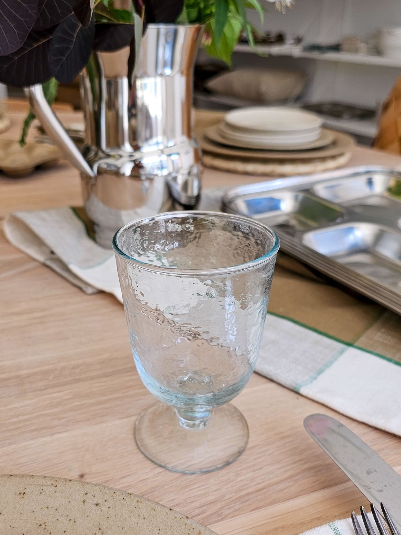 A clear textured glass goblet with a footed base, shown on a wooden table in a styled dining setting with silverware, plates, and a metallic pitcher in the background.