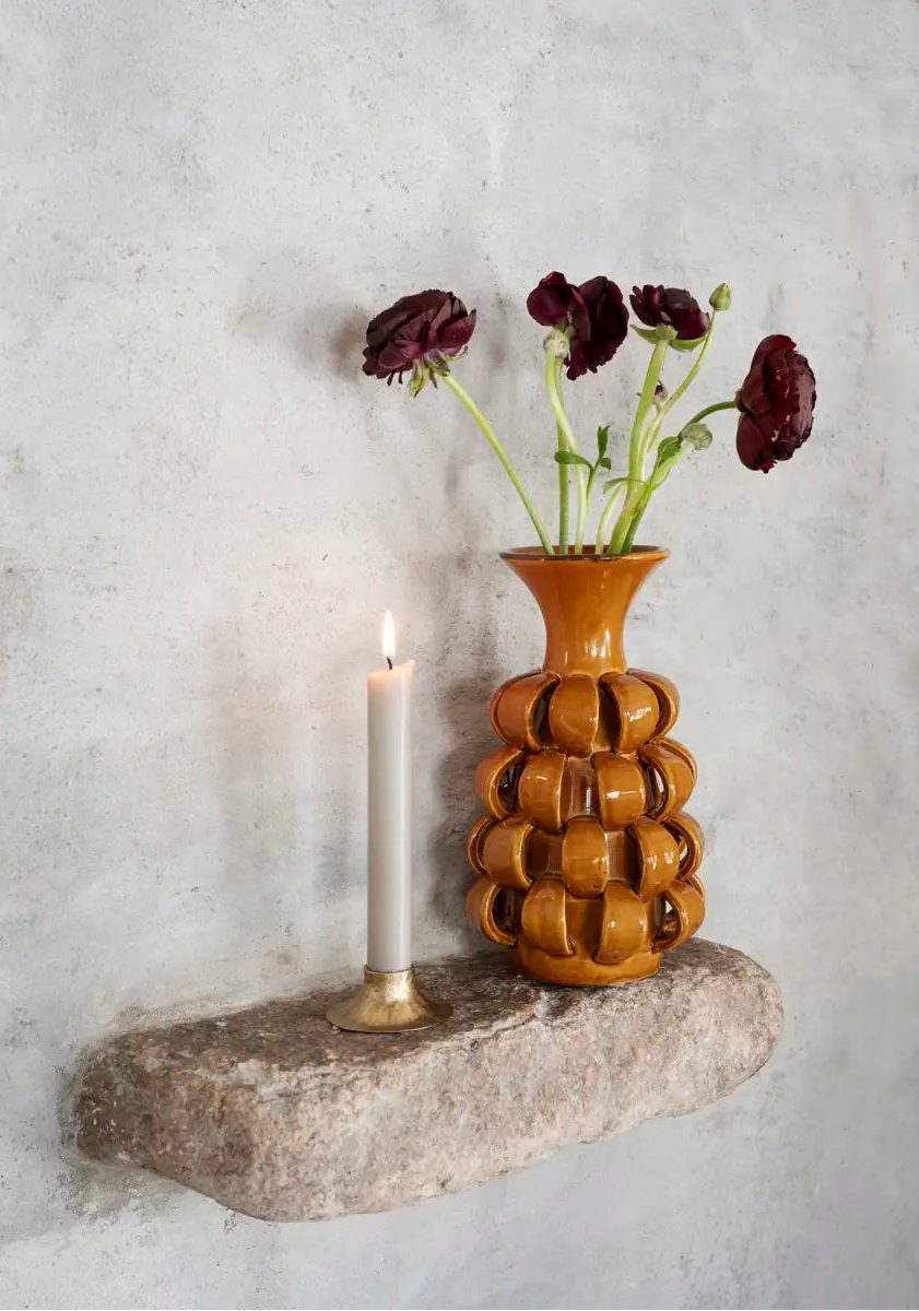 An amber-brown ceramic vase with a textured, bubble-like pattern sits on a stone shelf next to a lit white candle. The vase holds dark burgundy flowers and is set against a textured gray plaster wall.