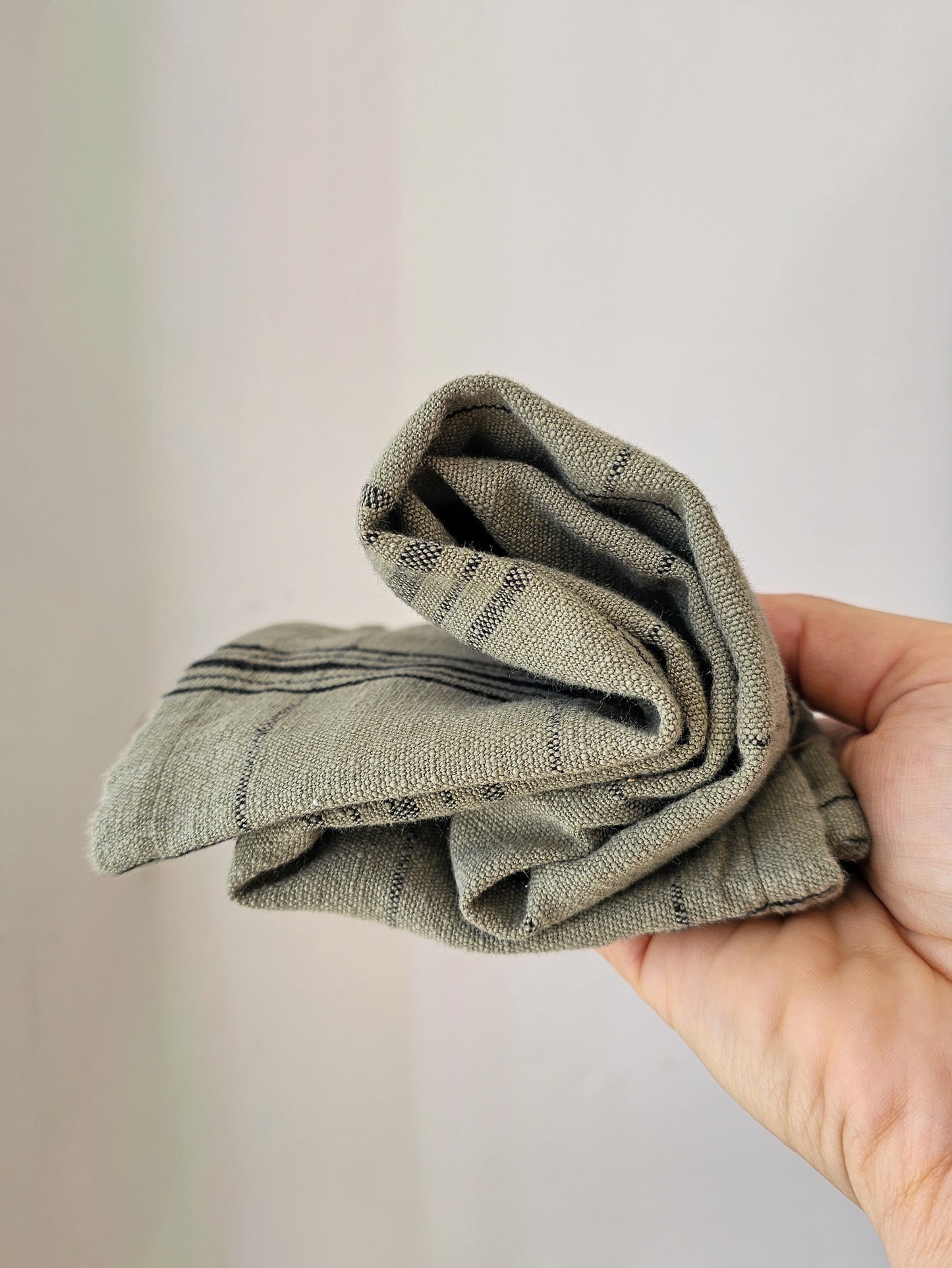 A hand holding a folded, textured cotton tea towel in a muted sage green color with subtle dark stripes, set against a soft, light beige background.