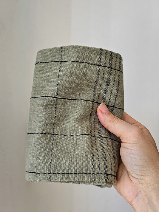 A hand holding a folded tea towel, likely in a muted green color with a dark windowpane plaid pattern, set against a plain off-white background.