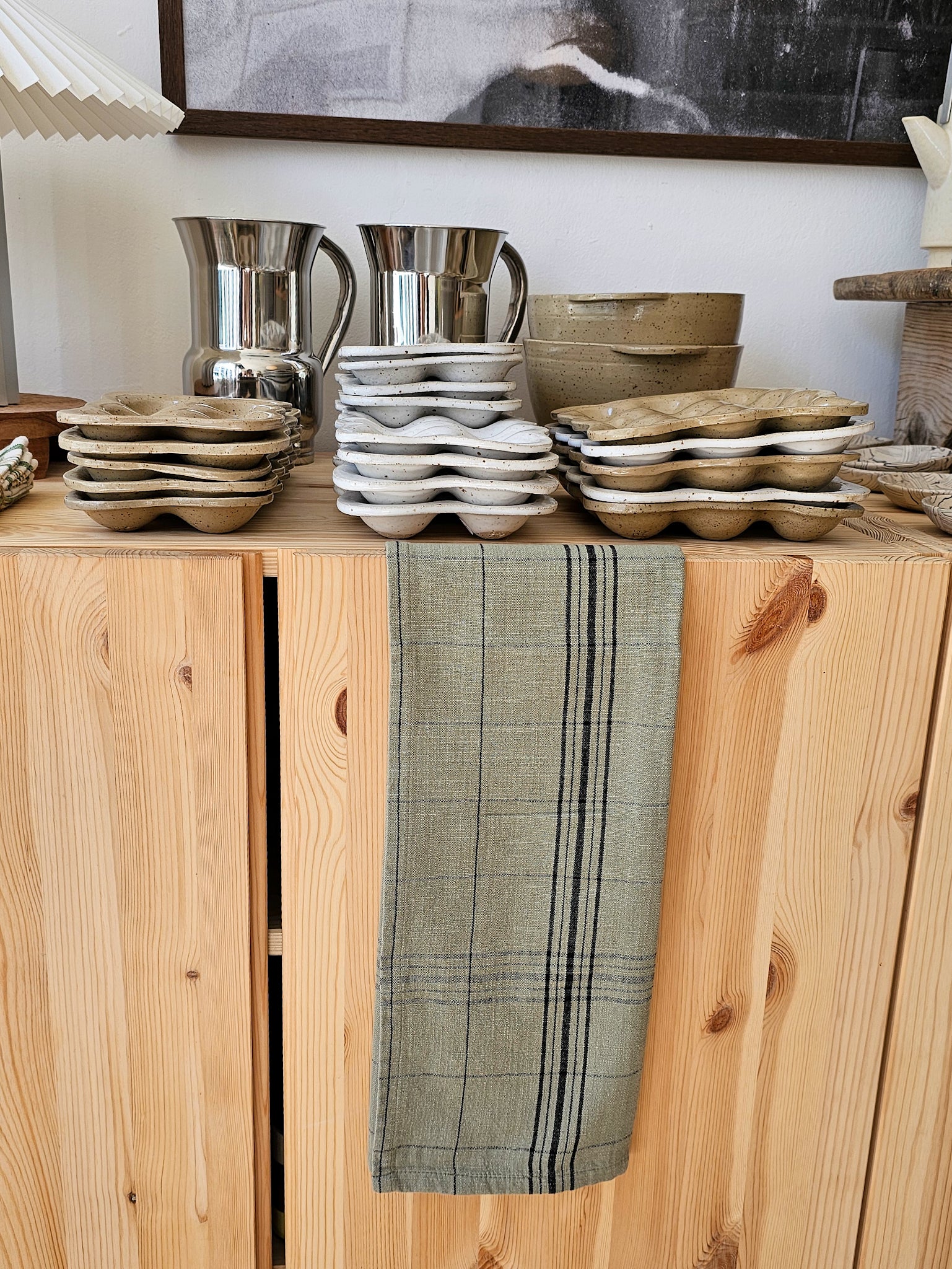 A kitchen scene showing stacks of ceramic egg-shaped dishes and two stainless steel pitchers on a light wood cabinet, with a green plaid towel hanging from the front door.