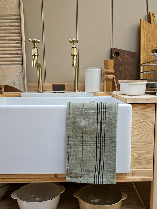A white farmhouse-style sink with two brass faucets, set in a rustic kitchen with light wood cabinetry. A sage green striped towel is draped over the front edge of the sink.