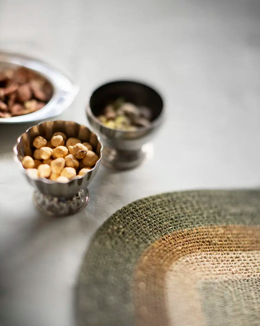 A close-up, shallow depth-of-field shot of a woven placemat with concentric arches in olive green, tan, and brown, with small steel ice cups of nuts blurred in the background on a gray surface.