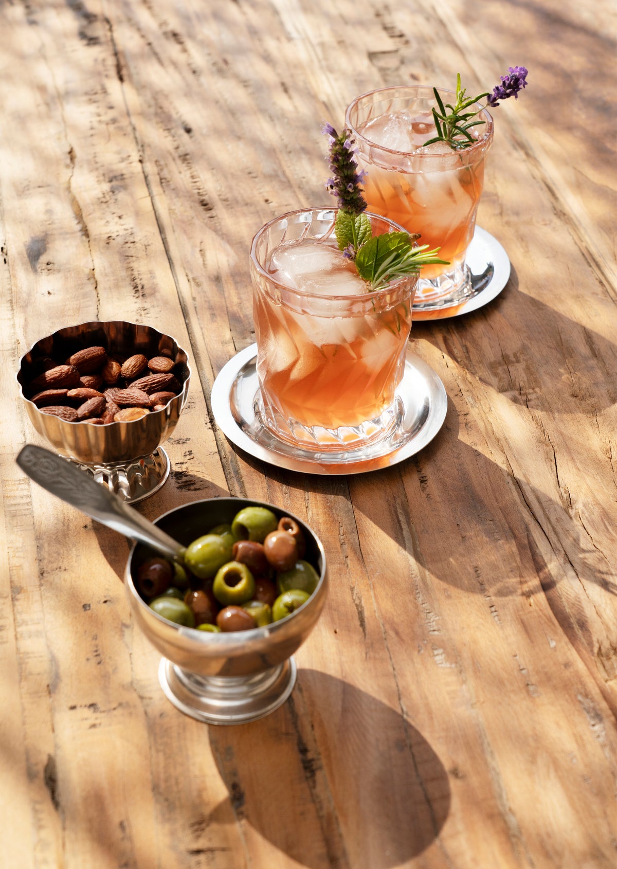 Two cocktails with ice and garnishes on a wooden table, accompanied by small steel bowls of olives and nuts.