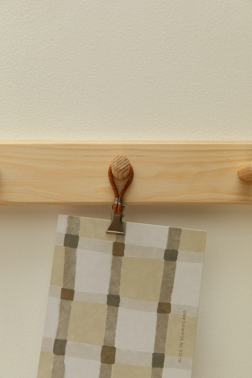 Close-up of Meraki towel clip in silver and leather hanging an Alice in Scandiland grid check art card from a pine shaker peg rail.