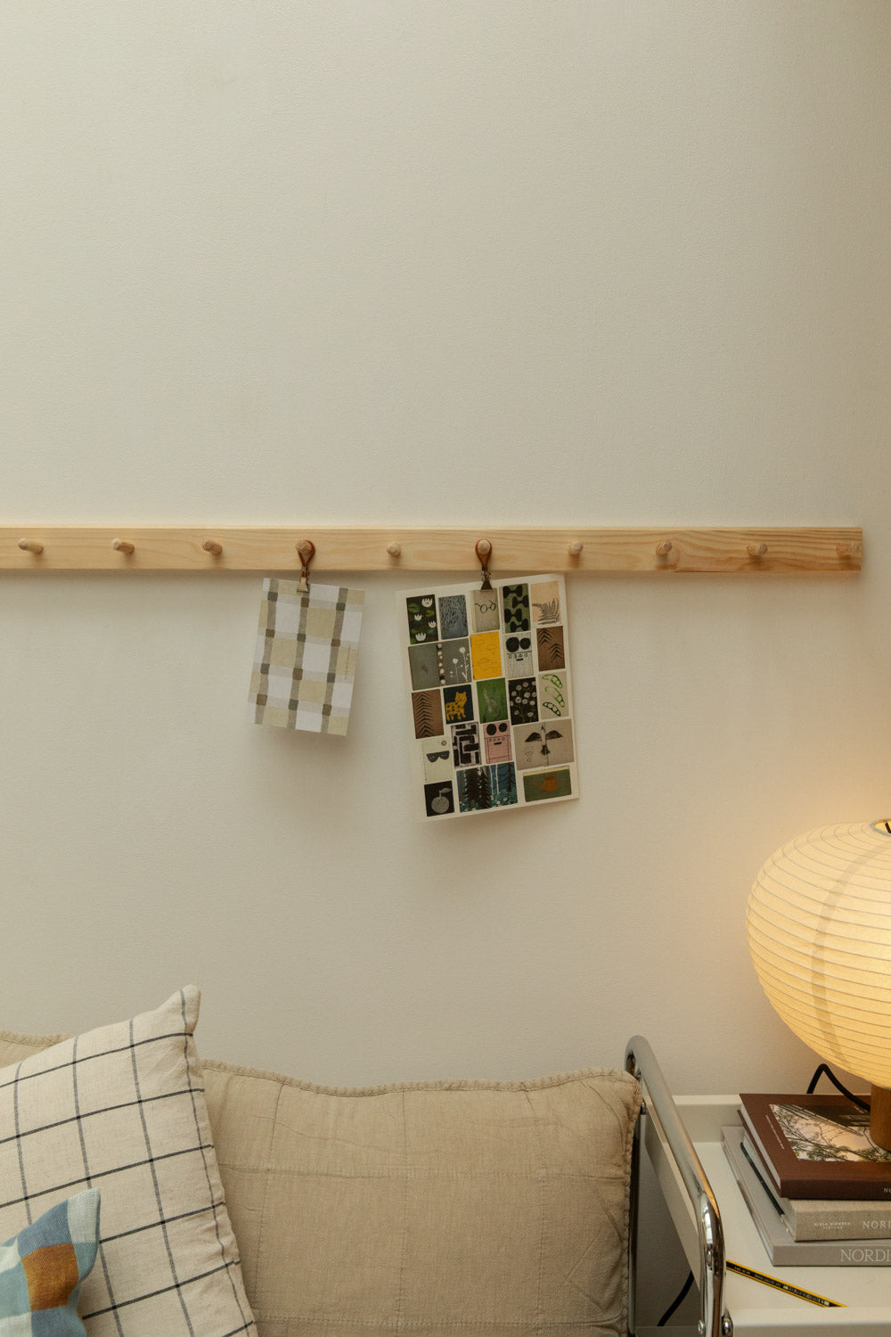 Two Meraki towel clips in silver and leather displaying an Alice in Scandiland grid check card and botanical collage card on a pine shaker peg rail above a linen sofa with grid check cushion and glowing paper lantern table lamp.