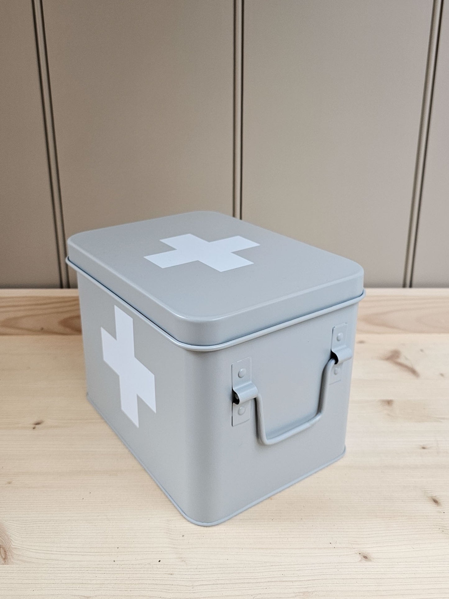 A light gray metal first aid box with white cross symbols on the lid and side, featuring a built-in handle, placed on a light wood surface against a paneled beige wall.