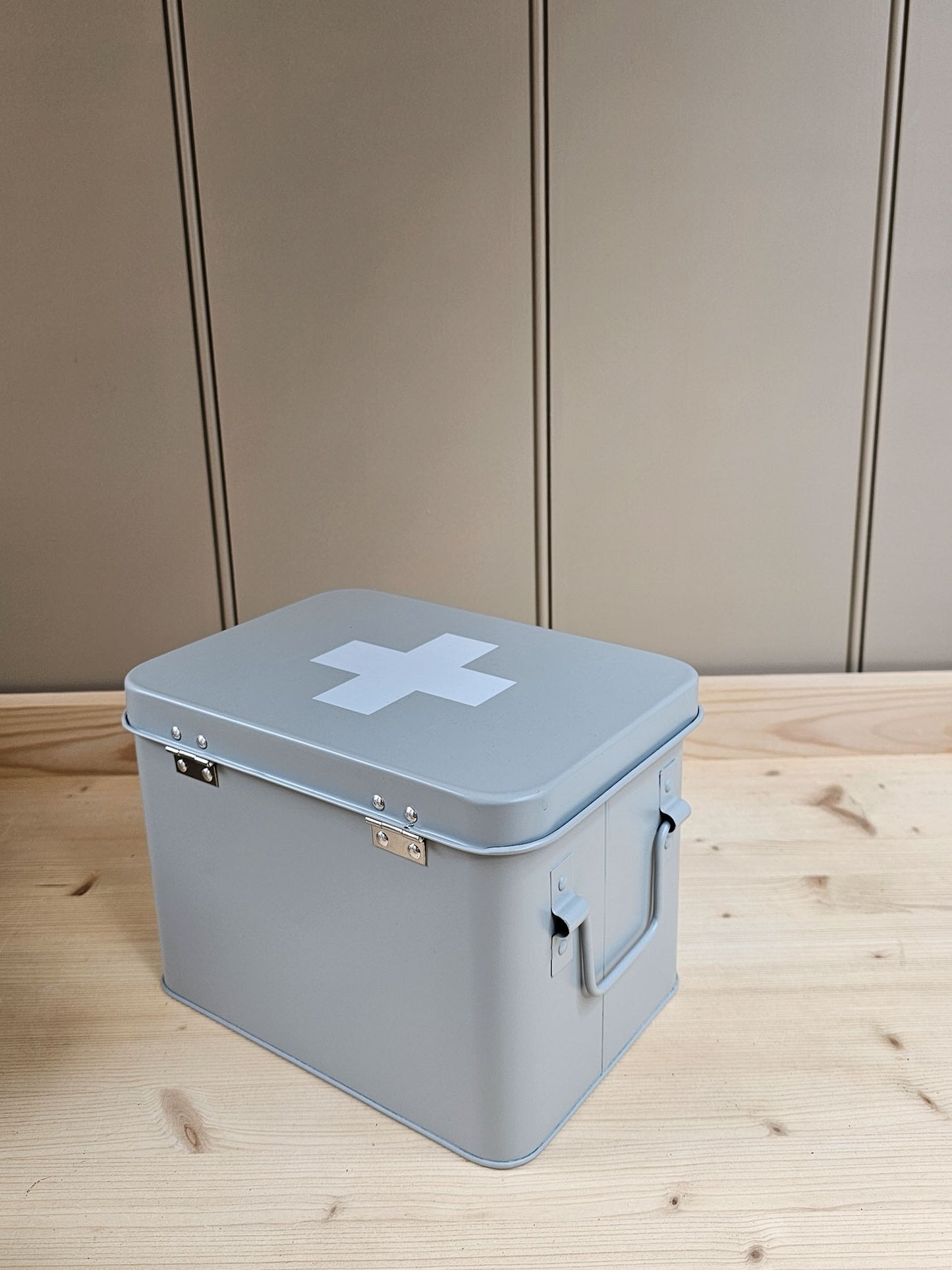A light gray metal storage box with a white cross symbol on the lid, placed on a light wood surface against a beige paneled wall.