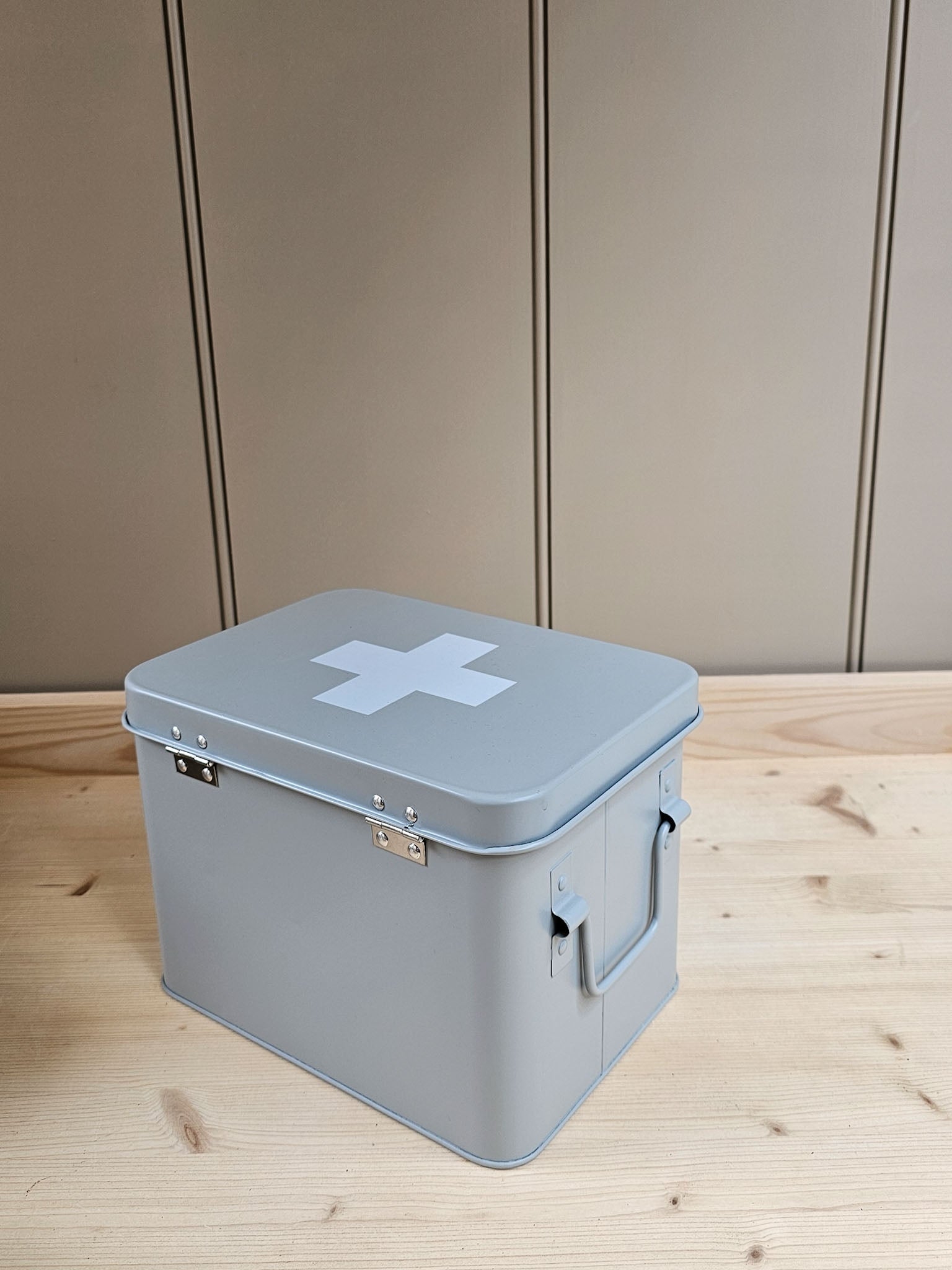 A light gray metal storage box with a white cross symbol on the lid, placed on a light wood surface against a beige paneled wall.