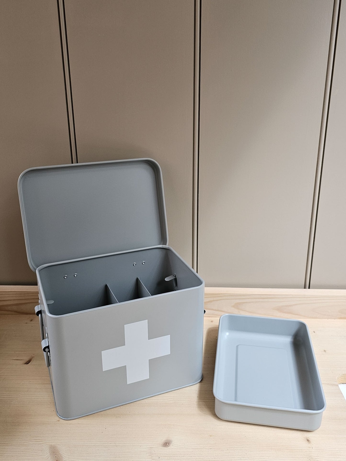 A light gray metal first aid box with an open lid, featuring a white medical cross on the front and two interior compartments. A separate shallow tray is placed beside it on a light wood surface against a beige paneled background.